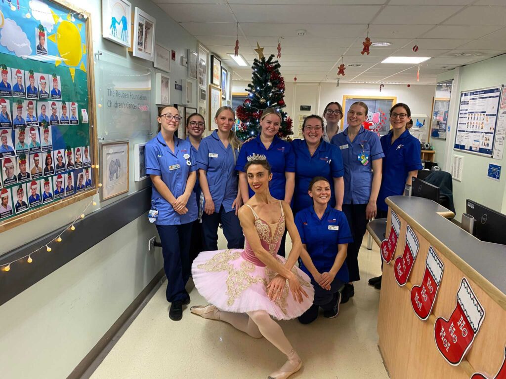 A smiling ballerina curtseying on a hospital ward, with a group of smiling female nurses.