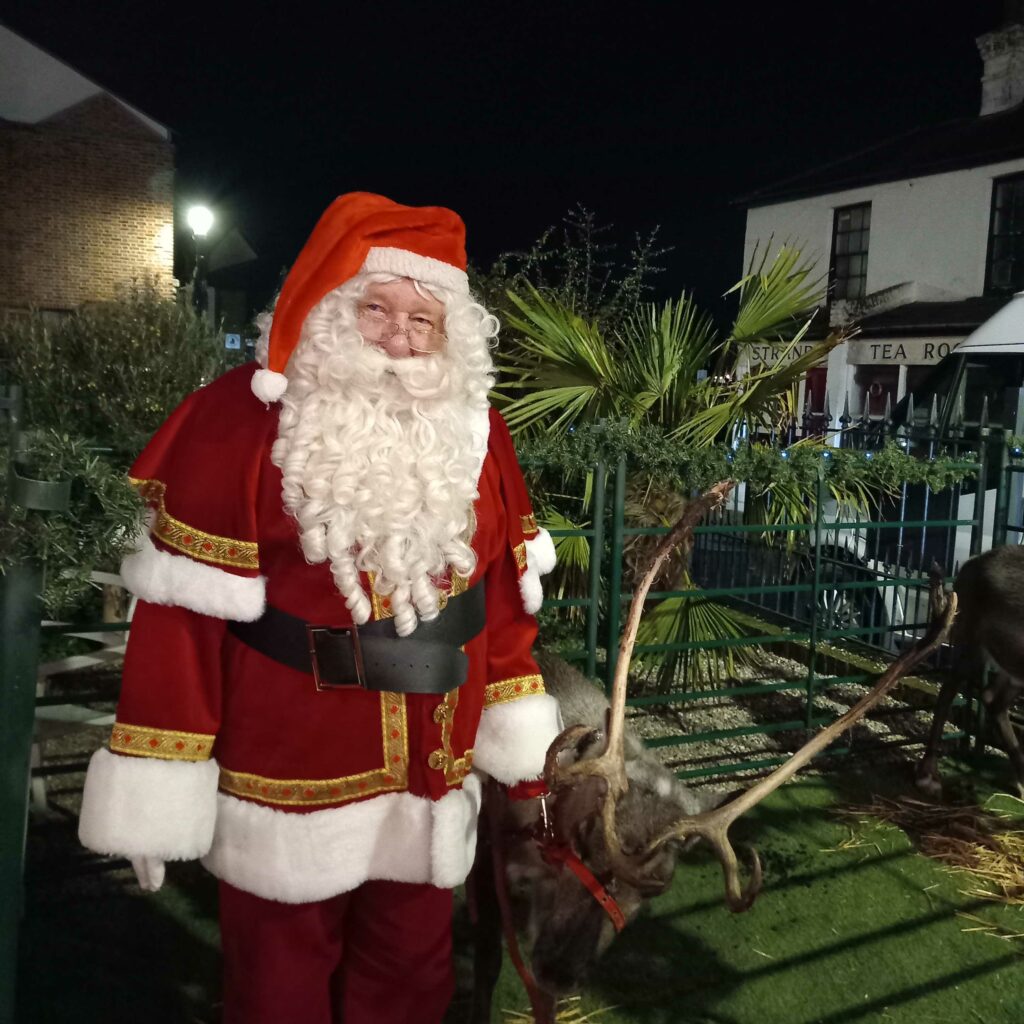 Father Christmas smiling and standing with a real reindeer outside the Gold Geese Grotto